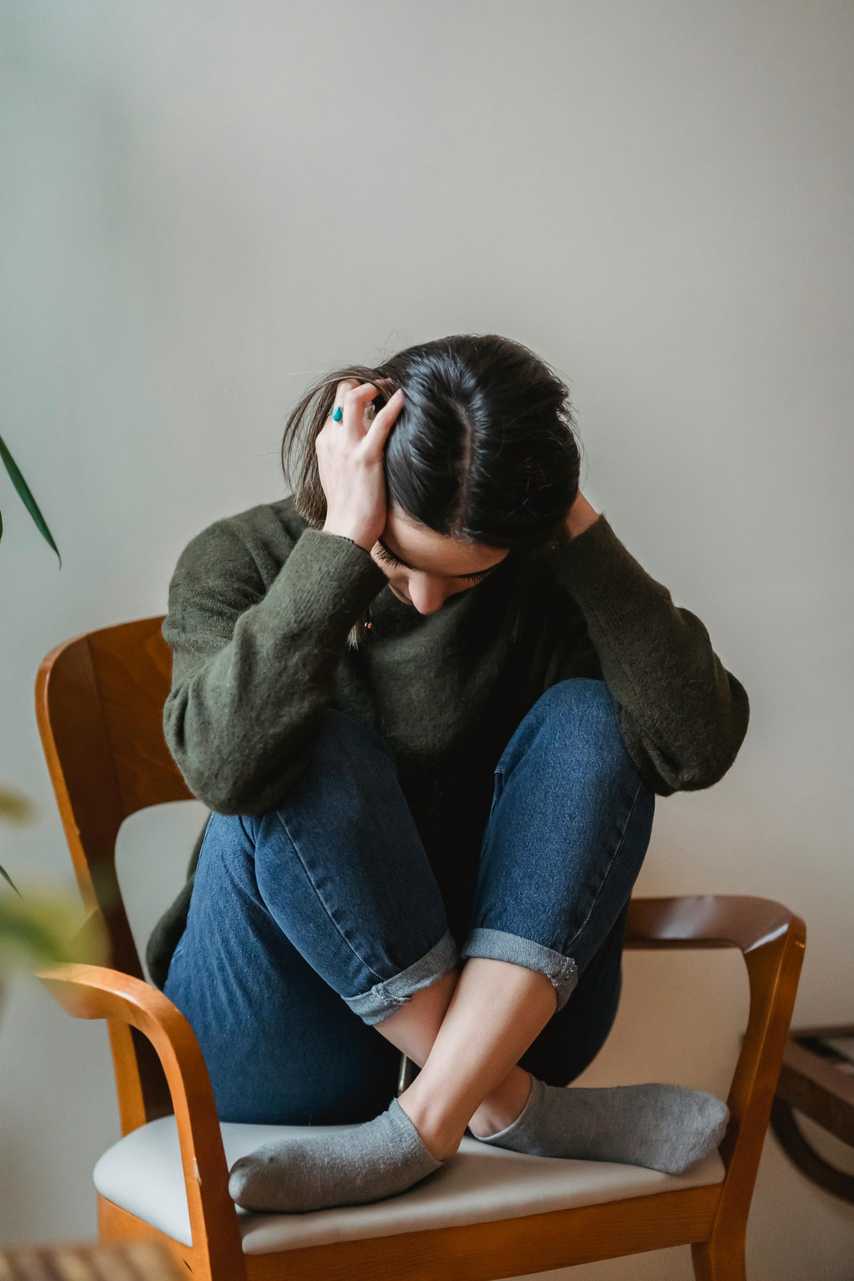 A young woman sitting on a chair indoors, holding her head in distress.